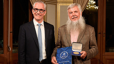 Der GI-Präsident Martin Wolf und der Preisträger Gerhard Weber bei der Preisverleihung der Klaus-Tschira-Medaille. Weber präsentiert die Urkunde und Medaille in die Kamera. Sie stehen in einem prunkvollem Raum mit einer gold-verziehrten Wand hinter ihnen