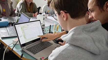 Ein jugendliches Kind sitzt am Laptop und bekommt etwas von einem erwachsenen Mann am Laptop gezeigt