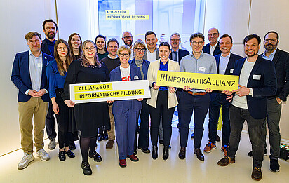 Auf dem Foto ist eine Gruppe von Menschen zu sehen. Sie halten Schilder hoch, auf denen steht: "Allianz für informatische Bildung" und "#InformatikAllianz"