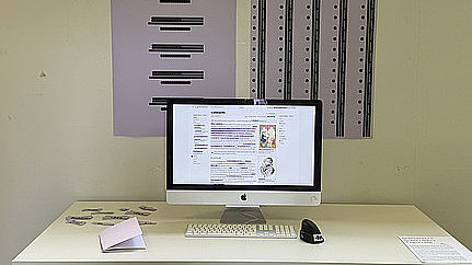 Ein Computerbildschirm mit schwarzer Maus und weißer Tastatur stehen auf einem Tisch. Daneben liegen fliederfarbene Papierschnipsel mit dem Aufdruck *in und *innen. Hinter dem Bildschirm hängen zwei große Plakate an der Wand auf welchem schwarze Punkte und Striche in regelmäßigen Abständen aufgedruckt sind.