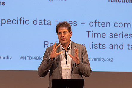 Prof. Glöckner während einer Präsentation auf einer Konferenz, hinter ihm sind Folien an eine Leinwand projeziert
