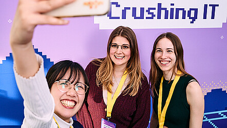 Drei Studierende machen ein Selfie vor einer Pressewand: Tech Crush – Crushing IT