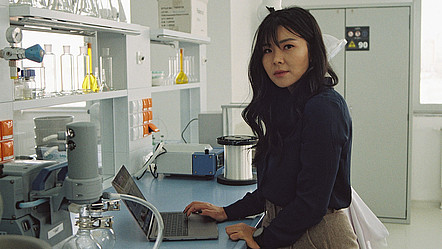 Eine junge Frau steht in einem Labor vor einem aufgeklappten Laptop, auf einem Regal stehen mehrere Glascontainer und Forschungsgeräte