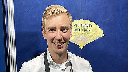 Portrait of Mattis Hartwig infront of the GI-Booth at IJCAI. He ist wearing a white shirt and smiling to the camera.