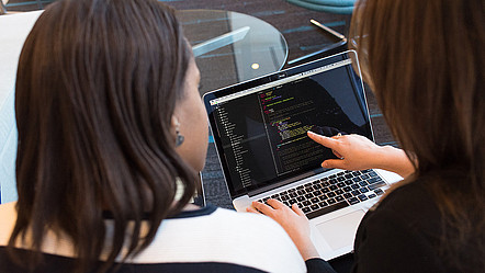 Zwei Frauen vor einem Laptop mit Programmiercode, eine von ihnen zeigt auf den Bildschirm
