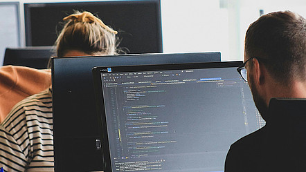 Zwei Personen arbeiten an Computerbildschirmen, auf einem Monitor ist Programmcode zu sehen.