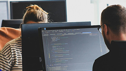 Zwei Personen arbeiten an Computerbildschirmen, auf einem Monitor ist Programmcode zu sehen.