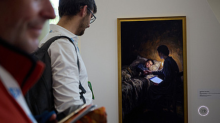 Ein Exponat der Ausstellung: Ein Screen in einem historisch wirkenden Bilderrahmen, erst auf den zweiten Blick ist erkennbar, dass darin kein Gemälde ist. Zu sehen ist eine Person, die mit einem iPad vor einem Bett sitzt, in dem ein krankes Kind liegt. Die Szene wirkt wie ein altes Gemälde, ist aber KI-generiert.