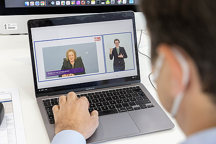 Eine Person sitzt vor einem Laptop. Auf dem Display ist ein Livestream des KI-Camps 2021 zu sehen, der simultan in Gebärdensprache übesetzt wird.