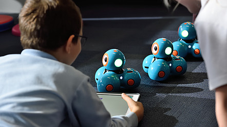 Ein kleiner Junge mit blauem Hemd, braunen Haaren und einer Brille steuert drei kleine blaue Roboter mit einem Tablet.