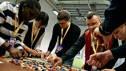 Eine diverse Gruppe junger Menschen bei einem Workshop, sie bauen Prototypen mit bunten Legosteinen.