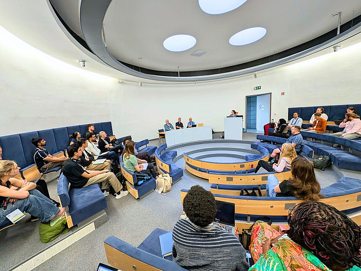 Blick in einen runden Hörsaal mit mehreren Sitzreihen. Rund 30 Personen sitzen im Halbkreis und verfolgen eine Veranstaltung mit einem Redner am Pult und drei Personen an einem Tisch.