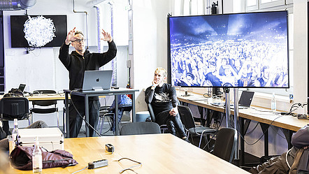 Ein Speaker des Workshops zeigt den Teilnehmenden eine Szene aus einem Konzert, in dem das Publikum Smartphones in die Luft hält.