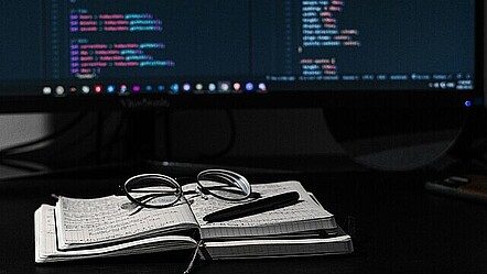 Ein Notizbuch, auf dem eine Brille und ein Stift liegen, aufgeschlagen vor einem Bildschirm, der Programmiercode zeigt