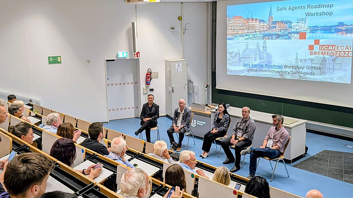 ALT-Text: Blick in einen Hörsaal während des „Safe Agents Roadmap Workshop“ auf der IJCAI-ECAI 2026 in Bremen. Fünf Personen sitzen bei einer Podiumsdiskussion vor dem Publikum, im Hintergrund ist die Präsentationsfolie des Workshops zu sehen.