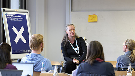 Ulrike Lucke sitzt auf einem Schreibtisch in einem Workshop-Raum und unterhält sich mit anderen Teilnehmenden. Hinter ihr ein Poster: NFDIxCS