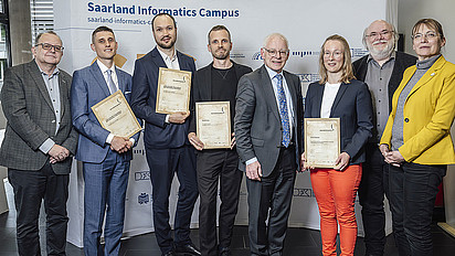 Acht Menschen vor einer Wand mit der Aufschrift "Saarland Informatics Campus". Vier der Acht präsentieren ein Zertifikat mit der Aufschrift "Journalismuspreis Informatik 2025" in die Kamera.