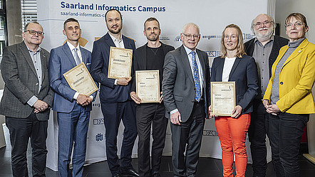 Acht Menschen vor einer Wand mit der Aufschrift "Saarland Informatics Campus". Vier der Acht präsentieren ein Zertifikat mit der Aufschrift "Journalismuspreis Informatik 2025" in die Kamera.