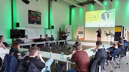 Blick in den Saal, in dem der Wettbewerb stattfand. Die Teilnehmenden sitzen um die Bühne, auf der Moderatorin Elena Müller gerade spricht.