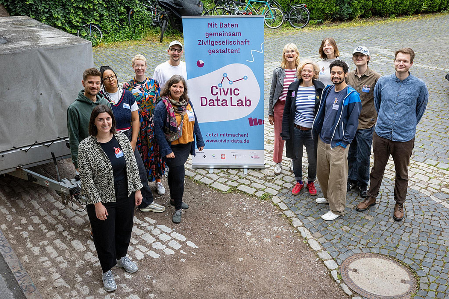 Teamfoto des Projekts Civic Data Lab: Zwölf Personen von oben fotografiert, sie lächeln und stehen neben einem Roll-up mit dem Logo des Projekts und dem Spruch: Mit Daten gemeinsam Zivilgesellschaft gestalten!