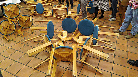 Auf dem Fliesenboden liegen mehrere Stühle ineinander verschränkt, rund um sie stehen Menschen und betrachten ihr Werk, das bei einer Team-Aufgabe passiert ist.