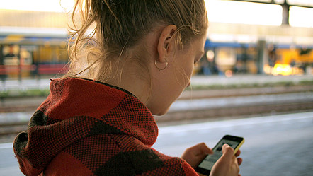 eine Frau die an einem Bahnhof steht und auf ihr Handy schaut