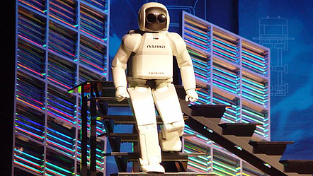 Zu sehen ist der humanoide Roboter ASIMO von Honda von einer Vorstellung 2004 an der Stanford Universität. Er geht eine Treppe auf der Bühne herunter und ähnelt mit seiner weißen Lackierung einem Astronauten.