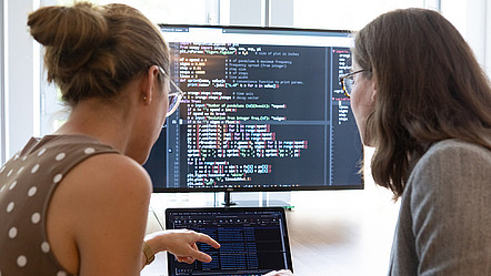 Zwei Frauen vor einem Laptop und einem Bildschirm, auf dem Code zu sehen ist