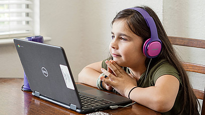Ein Mädchen mit Kopfhörern sitzt, das Kinn auf die Hände gestützt, vor einem aufgeklappten Laptop. 