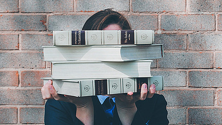 Eine junge Frau in einem dunkelblauen Hemd verbirgt ihr Gesicht hinter einem Stapel Bücher.