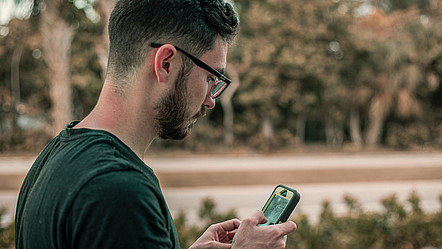 Ein junger Mann ist draußen unterwegs und benutzt währenddessen sein Smartphone.