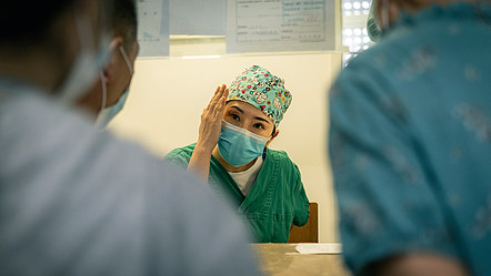 Eine Krankenhausangestellte im Gespräch mit zwei weiteren, sie trägt eine Maske und eine OP-Haube.