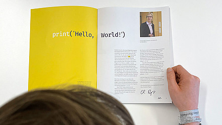 Eine Person blättert im aufgeschlagenen GI-Mitgliedermagazin. Zu sehen ist der Schriftzug: Hello, world!