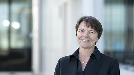 Claudia Eckert blickt lächelnd in die Kamera. Sie steht vor einem Gebäude mit Fenstern.