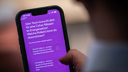Auf einem Smartphonebildschirm wird eine Frage an einen Spielteilnehmer gerichtet: "Dein Team braucht dich für eine Cybermission im Energiesektor! Welche Rolle(n) kannst du übernehmen?"