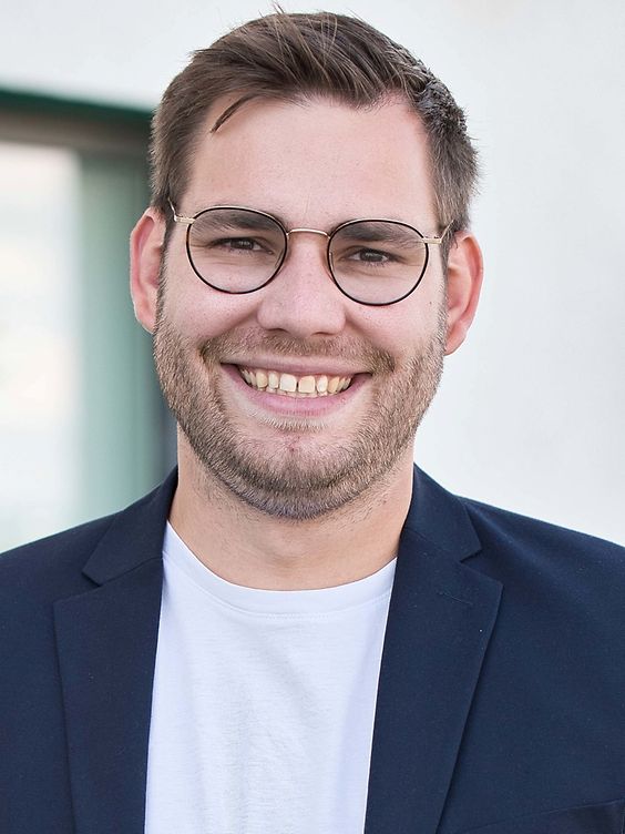 Porträt eines lächelnden Mannes mit Brille, Bart und kurzem Haar, in einem dunklen Blazer und weißem T-Shirt, vor einem hellen Hintergrund.