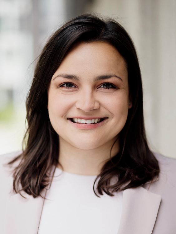 Porträt einer lächelnden Person mit schulterlangem dunklem Haar in einem hellen Blazer, aufgenommen vor einem unscharfen Hintergrund.